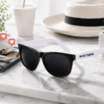 Custom promotional sunglasses with logo imprint styled next to a phone displaying Instagram likes and photos